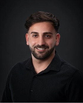 Homme brun jeune et barbu habillé en noir sur fond noir.